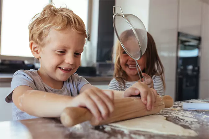 Two happy kids are baking and rolling out dough on a wooden countertop.
