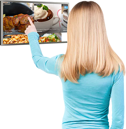 A woman pointing at the screen of a brushed stainless steel kitchen TV on a white wall.